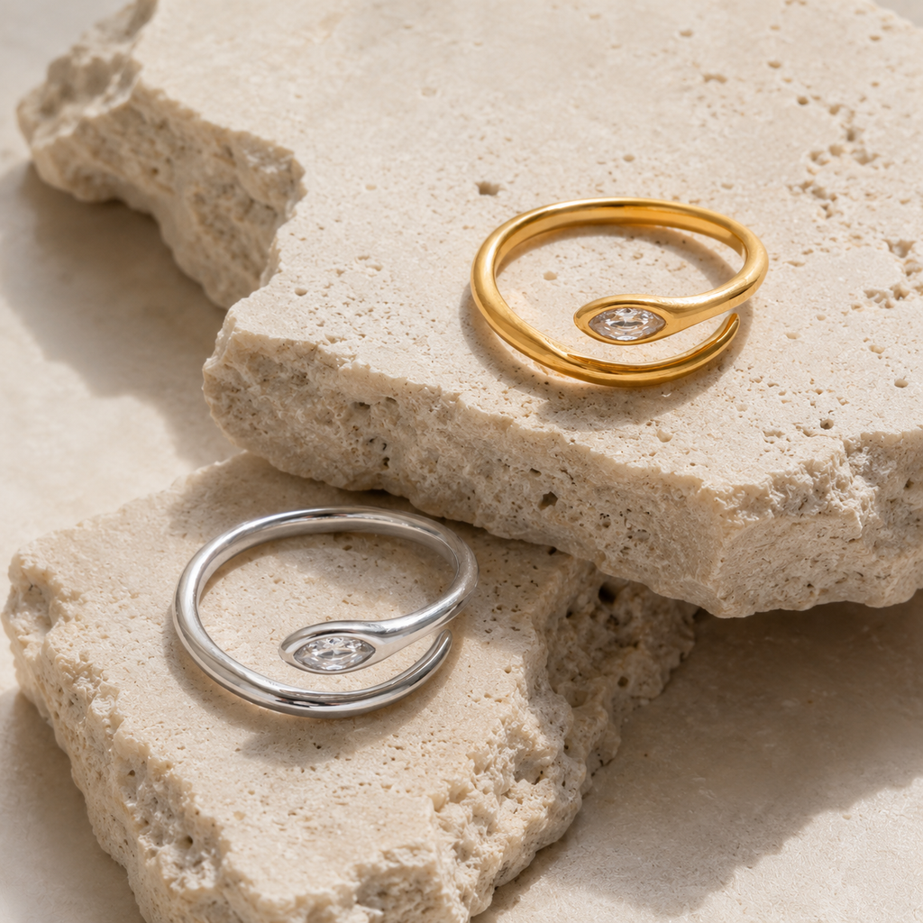 Gold and silver adjustable open-band rings with marquise crystal detail displayed on raw travertine stone.
